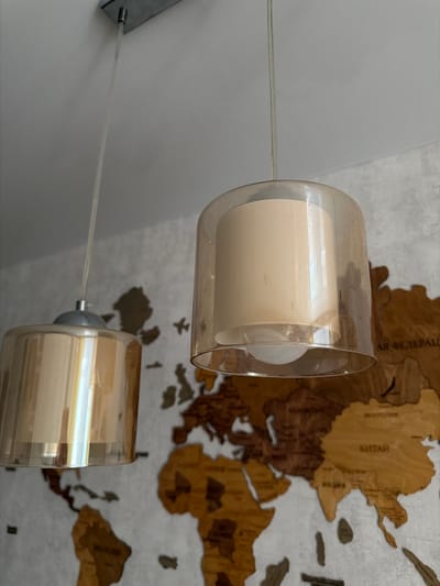 A close-up, eye-level shot shows two pendant light fixtures hanging from a white ceiling. The fixtures are cylindrical, with a translucent, amber-colored glass shade and an inner cream-colored shade. The left fixture is closer to the camera and appears to be slightly shorter. The right fixture is further away and slightly taller. The lights are illuminated by what appear to be standard light bulbs.
Behind the light fixtures, a large, wooden world map is mounted on a textured, white wall. The map is composed of different wood tones, highlighting continents and countries. Some labels in a non-English script are visible on the map, particularly in Asia. The lighting in the image is soft and diffused, casting subtle reflections on the glass shades of the light fixtures. The scene is indoor, likely a room in a residential or commercial space. The specific location in Semiluki, Russia, cannot be determined from the image itself.