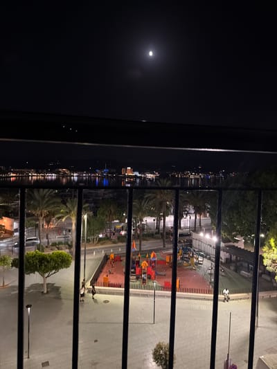 A nighttime aerial view captures a waterfront scene in Sant Antoni De Portmany, Spain. The image is framed by a dark railing in the foreground. Beyond the railing, a brightly lit cityscape with numerous buildings and lights reflects on the calm water of the bay. A full moon is visible in the dark sky. Below the cityscape, a playground area with colorful equipment is illuminated. Palm trees and streetlights dot the area surrounding the playground. Several people are visible walking and sitting in the illuminated public space. A car is parked on the left side of the image. The ground is paved with patterned tiles.