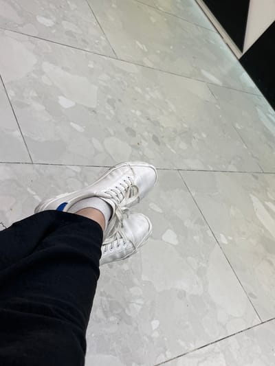 The image shows a close-up view of a person's legs and feet, with white sneakers and black pants, resting on a light gray tiled floor. The tiles have a marbled pattern with various shades of gray and white. In the upper right corner of the image, a portion of a black and white checkered pattern is visible, possibly part of a wall or structure. The lighting suggests an indoor environment. The location in Aktau, Kazakhstan, is not evident from the image itself, as there are no identifying landmarks or context clues.