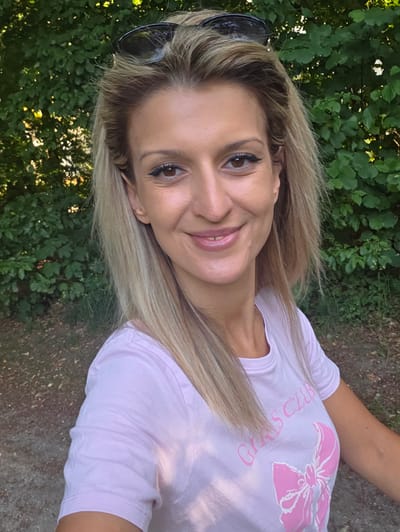 A blonde woman with her hair down and sunglasses on her head smiles at the camera. She is wearing a pink t-shirt with a bow graphic and the words "GIRLS CLUB" printed on it in pink lettering. The background is a blur of green foliage and brown ground, suggesting an outdoor setting, possibly a park. There are no discernible landmarks to indicate a specific location in Munich, Germany.