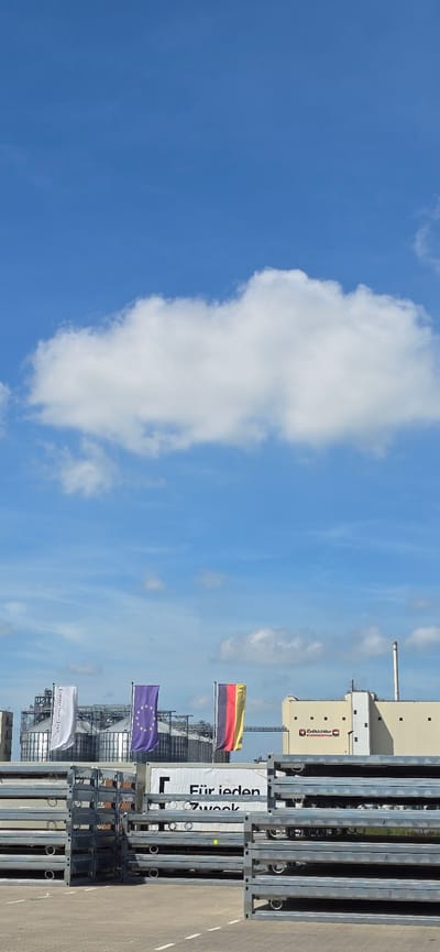 The image shows an outdoor scene with a clear blue sky and scattered white clouds. In the foreground, stacks of grey metal frames are neatly arranged on a paved surface. Behind the frames, there are three flags displayed vertically on poles: a white flag with black text, a purple flag with yellow stars (likely the European Union flag), and a flag with horizontal stripes of black, red, and yellow (the German flag). Further in the background, industrial buildings are visible, including large silos and a light-colored building with a smokestack. The text "Reth'Hötter" and "Industriestr. 33. 49733 Haren (Ems)" is visible on a logo on the light-colored building, indicating the location as Haren (Ems), Germany. There are no people visible in the image.