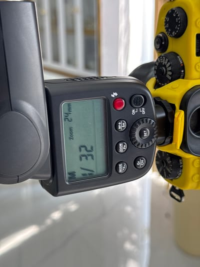 This close-up media file features a camera flash unit mounted on a yellow camera body. The flash unit has a digital display showing "M V32" and "Zoom 24mm". Various buttons are visible on the flash unit, including "SET", "ON/OFF", "CH/Gr", "ZOOM", "MF", and "MODE". The camera body has several dials, one of which is labeled "MOVIE" and another with settings like "60", "125", "250x", "500", "1000", "2000", "4000", and "8000".

The scene appears to be an indoor setting, possibly a studio or a room, with natural light casting shadows on a white surface in the background. There are no people visible in the media file. The focus is entirely on the camera and flash equipment. The overall impression is one of technical equipment, possibly in use for photography or videography, with the digital display suggesting settings for aperture, shutter speed, or flash power. The presence of the "MOVIE" label on the camera dial might indicate it is a camera with both still photography and video capabilities. The sunlight casting shadows suggests it might be daytime, but no specific time of day can be determined. No other location cues are available.