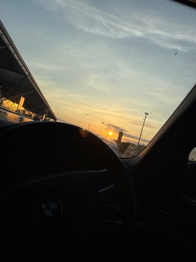Here's a description of the image:
The image is taken from inside a car, looking out through the windshield at a sunrise or sunset. The sun is positioned low on the horizon, casting an orange glow across the sky. Below the horizon are the structures of an airport, possibly an aerial tramway. A BMW steering wheel is visible in the foreground on the right side of the image. The rest of the dashboard and interior of the car are dark. The windshield shows some minor spotting. While the image includes the BMW logo, I cannot identify the specific location within Uttlesford, United Kingdom with the available information.