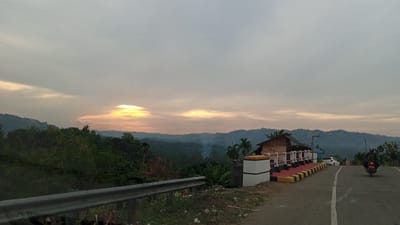 The image captures a scenic view of Buketmulia, Indonesia, likely during the early morning or late evening, indicated by the soft, diffused sunlight breaking through an overcast sky. The foreground is dominated by a paved road curving to the right, with a metal guardrail on the left. Along the right side of the road, a small, rustic wooden structure with a corrugated metal roof is visible, adorned with a white and red painted railing. A white car is parked near the structure. Further along the road, a person is riding a motorcycle away from the viewer. The mid-ground features a lush, verdant landscape of rolling hills covered in dense vegetation, including palm trees and what appears to be agricultural crops. Wisps of smoke rise from the distant hills, suggesting ongoing agricultural activity or a controlled burn. The background is characterized by a series of layered, misty mountains fading into the horizon under a pale, cloudy sky.