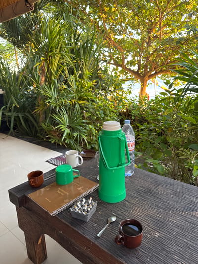 The image depicts an outdoor table set in what appears to be a tropical setting, possibly a veranda or cafe overlooking a body of water. The table is made of dark, weathered wood and is laden with various items. In the foreground, to the left, is a small brown mug. Next to it is a brown notebook with a spiral binding, topped with a green plastic lid. To the right of the notebook is a grey ashtray filled with cigarette butts. Further to the right, a silver spoon rests near a small brown mug filled with a dark liquid, likely coffee. Behind these items, a tall, bright green thermos with a white lid and handle stands prominently. Next to the thermos is a clear plastic bottle of water. A white ceramic mug sits to the left of the green thermos.

The background is dominated by lush green foliage, suggesting a location rich in vegetation. Sunlight filters through the leaves, creating dappled light. In the distance, a hint of a beach and calm water can be seen, with colorful flags or banners visible, suggesting a public area or resort. The overall impression is one of a relaxed, possibly morning or late afternoon setting, conducive to enjoying a beverage and a cigarette while taking in the natural surroundings. The mention of Abang, Indonesia, places this scene in a specific geographical context known for its tropical climate and coastal beauty.