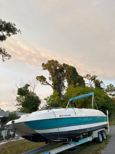A white and teal speed boat is parked on a trailer on a grassy area next to a paved road. The boat has a blue bimini top and the license plate "FL 6926 HB" is visible on the side of the boat. Behind the boat and trailer are dense green trees and foliage, with a house visible in the background to the left. The sky above is cloudy with hints of pink and orange, suggesting it is either sunrise or sunset. The scene is set in South Venice, Florida, United States of America.
