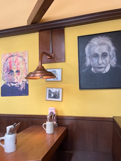 The image depicts a cafe interior in Wien, Austria at coordinates 48.212 latitude and 16.359 longitude. The cafe is brightly lit with yellow walls and wood paneling. A wooden table is in the center of the image, with two beer steins filled with cutlery on it. There is a light fixture mounted on the wall behind the table, consisting of a copper lampshade and an arm extending from the wall. Above the table, to the left, is a framed, colorful portrait of Albert Einstein. To the right of the table, a larger framed black and white portrait of Albert Einstein hangs on the wall. Above the larger portrait, there is a framed black and white photo of a street sign. The space is a cafe, or possibly a bar, with seating at a wooden bench along the wall. The cafe is likely a place for people to gather and socialize.  The image does not depict people in the scene.