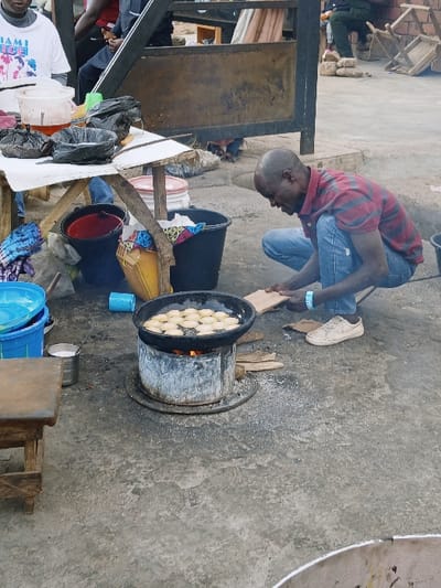 The image captures an outdoor scene, likely a street food stall or an informal market setup, where food is being prepared. The ground is a hard, grey surface, possibly concrete.
In the center of the frame, a man with a shaved head, wearing a red and black plaid collared shirt and blue jeans, is squatting down. He is focused on a large, dark pot filled with numerous round, light-colored food items, which appear to be frying or cooking. The pot is placed on a robust, cylindrical metal or clay stove, from which flames are visible, indicating that wood or charcoal is being used as fuel. The man holds a wooden stick or paddle, suggesting he is stirring or turning the food in the hot oil. He has a blue band around his left ankle, on which the word "OVER" is visible. White sneakers are placed on the ground next to him.
Around the cooking area, various containers and makeshift structures are present. To the left of the cooking pot, there is a large black bucket. Further to the left, behind the man and near a wooden support structure, another black bucket contains a reddish liquid, possibly oil or batter. Next to this, a blue plastic container is visible on the ground. A rustic wooden stand supports various items, including small plastic jugs (one white, one clear) and several black plastic bags that are either empty or contain goods. Partially visible in the bottom left corner is another person wearing a white t-shirt with graphics, where the word "MIAMI" is discernible. In the far background on the left, parts of another person and some wooden objects or furniture can be seen.
The scene depicts an active food preparation process, highlighting the use of traditional cooking methods in an outdoor setting. The overall atmosphere suggests a busy, functional, and somewhat rustic environment.