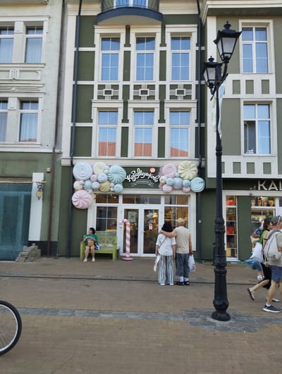 The image captures a street scene in Zelenogradsk, Russia, on a bright day. In the foreground, the wheel of a bicycle is visible on the left, suggesting it's parked or being ridden nearby. The main subject is a building with a green facade and large windows. A black lamppost stands prominently in front of the building.

The building appears to be a commercial establishment, possibly a shop or cafe, with a cheerful facade decorated with large, colorful meringue-like sculptures above the entrance. A sign with Cyrillic text, "Сладкоежка," is visible above the entrance, which translates to "Sweet Tooth." The entrance itself features glass doors. To the right of the entrance, a large pink and white candy cane decoration stands vertically.

Several people are present. A woman is sitting on a green bench in front of the building, holding a bag of snacks. Two people, likely a couple, stand with their arms around each other, looking towards the building. Further to the right, a group of people are walking past, some carrying shopping bags, and one person is running. The people are dressed in casual summer clothing.

The setting is a paved walkway or street lined with buildings. The architectural style of the building suggests it's in a historic or quaint area. The sunlight indicates it's daytime, likely late afternoon or early evening given the long shadows cast by the lamppost and people. The overall mood is lively and inviting, with the colorful decorations adding a playful touch to the urban environment.