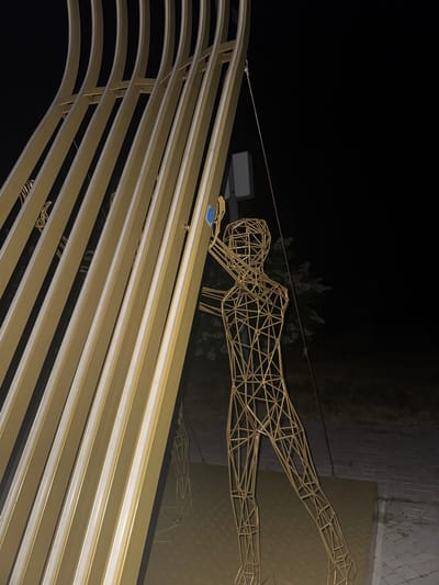 The image shows a golden, abstract sculpture resembling a human figure made of wireframe, standing in front of a large, curved, golden structure with vertical ribs. The scene is illuminated at night, with the background being dark, suggesting an outdoor environment. A small blue rectangular object is attached to the golden structure near the top of the wireframe figure. The ground appears to be paved with textured tiles. The location is identified as Egorlykskaya, Russia.