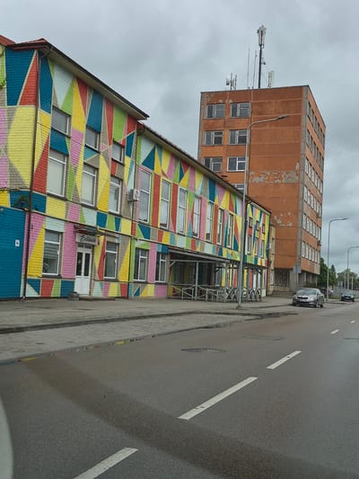 The image depicts a street scene in Šilutė, Lithuania, featuring a colorful, geometrically patterned building on the left and a tall, orange, weathered building on the right. The foreground shows a wet asphalt road with white lane markings and a sidewalk made of interlocking pavers. A silver sedan is parked on the side of the road in front of the orange building. The sky is overcast.
The colorful building, likely a commercial establishment, has a vibrant facade painted with large, overlapping diamond shapes in various bright colors, including yellow, pink, green, blue, and red, against a brick background. Several windows are visible on this building, along with a white entrance door. A sign above the door reads "Baldinake". The orange building, taller and older, appears to be an office building or apartment block, with numerous windows and a rough, peeling facade. It is topped with telecommunications equipment. The street is lined with lampposts, and there are some outdoor seating areas with tables and chairs visible in front of the colorful building. The overall impression is one of urban development with a juxtaposition of modern art and older architecture.