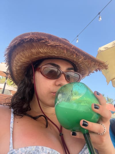 A person wearing a straw hat and sunglasses is drinking from a large green glass with a black straw. The person has dark hair, and dark nail polish. They are wearing a floral-patterned bathing suit and a ring on their finger. The background includes a clear blue sky, string lights, and partial views of umbrellas and palm trees. The scene appears to be in an outdoor setting, possibly a beach or patio area. While I cannot confirm the location specifically as Brisas de Zicatela, Mexico, based on the available image features, it could plausibly be a coastal resort location.