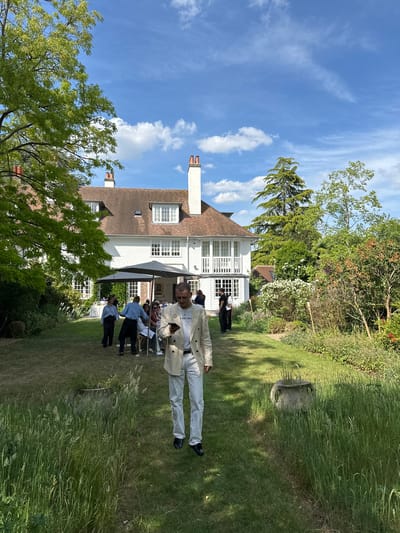 The image depicts a gathering in the garden of a white, two-story house, possibly located in Cambridge, United Kingdom. The weather is sunny with a blue sky and scattered clouds.
A man dressed in a light-colored suit stands in the foreground, partially obscuring a group of people seated at tables under umbrellas. He is holding a bag. Several other people can be seen, some dressed in light blue shirts and dark pants.
The backdrop features a well-maintained lawn, mature trees, and various plants. A stone garden ornament sits among the tall grass on the right side of the frame. The overall impression is that of a social event or party taking place in a residential garden.