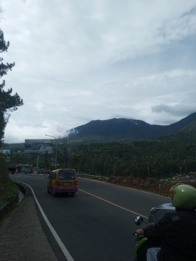 The image depicts a mountainous road scene in the city of Sandamal Eliya, Sri Lanka, under an overcast sky. In the foreground, the back of a person wearing a bright lime-green full-face helmet and a dark jacket is visible, operating a green scooter or small motorcycle. The vehicle is positioned on the right side of a two-lane asphalt road, which features a solid white line on the shoulder and a broken yellow center line, curving slightly to the left. Further down the road, a yellow and purple minivan drives away from the viewer.
To the left of the road, a steep, green, vegetated embankment with a concrete drainage ditch runs parallel to the asphalt. A tall conifer tree is partially visible at the far left edge. Adjacent to the road on the left, a large billboard with light blue supports displays an advertisement, partially reading "DPRK CLOUD". A street light pole stands near the billboard. Small, simple buildings and parked motorcycles are visible in the distance beyond the billboard, suggesting a roadside cluster.
The right side of the road shows a sloped area with disturbed brown earth and small, newly planted saplings, some with white protective wrapping around their bases. Multiple utility wires or power lines traverse the midground horizontally. In the background, dark green, densely forested mountains rise under the cloudy sky, with some peaks partially obscured by low-hanging mist or clouds. Distant residential structures are faintly visible at the base of the mountains on the right.