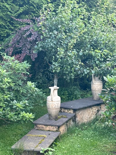 This is a medium shot, eye-level, vertical, outdoor photograph taken in Yukki, Russia. The scene depicts a garden with lush green foliage, including a large, dense tree in the center with abundant leaves. To the left of the tree, there is a bush with dark purple leaves. In front of the tree and to the right, there are two decorative stone urns. The urn on the left is closer to the viewer and features intricate carvings and a handle. The urn on the right is further away and also appears decorative. These urns are placed on a stone ledge which is supported by a stone wall. In front of the ledge, there are two stone steps descending into a grassy area covered with green grass and some moss. The lighting suggests it is daytime, possibly overcast or shaded, as there are no harsh shadows. The overall impression is one of a tranquil, cultivated garden space.