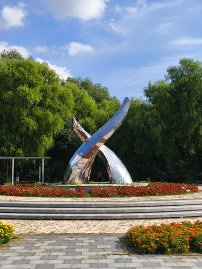 In Nanguan District, China, a large, metallic sculpture is the centerpiece of a public space. The sculpture, which appears to be made of polished steel, has a modern, abstract design resembling two intertwined or crossed elements that arc upwards. It is situated on a raised circular platform adorned with a vibrant bed of red flowers. Behind the sculpture, a dense backdrop of lush green trees creates a natural setting. In front of the platform, a set of tiered concrete steps leads down to a paved area. Further in the foreground, a patch of bright orange flowers is visible on the right, and a few yellow flowers are seen on the left, bordering the paved walkway. The sky above is a clear blue with scattered white clouds. A small, open-sided shelter with a light-colored roof stands to the left of the sculpture, and a person wearing a backpack is partially visible near the base of the sculpture.
