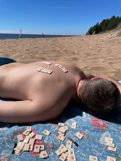 A man is lying face down on a beach in Zelenaya Roshcha, Russia, with Rummikub tiles on his back and scattered around him on a blue floral blanket. The beach has sand and scattered dry vegetation. In the background, the sea is visible under a clear blue sky. A windsurfer and other figures are in the distance. The man's back is exposed, and he appears to be sunbathing. The Rummikub tiles are various colors and numbers, suggesting a game is in progress or has been interrupted. The scene depicts a leisurely day at the beach.