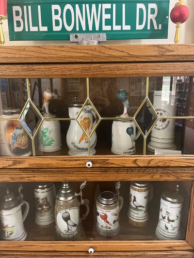 This interior image displays a wooden display cabinet featuring a collection of ceramic and stoneware steins, predominantly adorned with pigeon and fowl imagery, some bearing German text. Positioned above the cabinet is a green rectangular street sign with white lettering, clearly reading "BILL BONWELL DR." The sign is mounted on a light-colored wall using a metal bracket.

The cabinet, appearing to be made of oak, has glass doors with decorative leaded-glass patterns in a brass tone. The upper shelf contains various steins, including one with a standing figure, another with a prominent blue pigeon figure serving as a lid, and additional pieces with painted birds and German inscriptions such as "Erigerer Vollgucker" and "UILER KOPF." The lower shelf houses several traditional steins, most featuring hinged metallic lids and ornate handles. Designs on the lower steins include detailed pigeons, and text like "Zuchtschau 1987-G" and "Welt Schild" is visible. Small white circular knobs adorn the cabinet doors. Faint reflections in the glass panels suggest an indoor environment, with a partial reflection of a person in a white garment visible in the upper left section. A red spherical ornament with a gold base is partially visible on the right side of the frame, near the top.

The presence of the "BILL BONWELL DR." street sign, combined with the extensive collection of pigeon-themed steins, indicates the likely location is the Oklahoma City Pigeon Club, situated on Bill Bonwell Drive in Oklahoma City, United States. No direct human subjects or actions are depicted; the scene is a static display of collectibles.