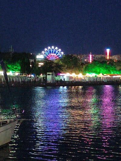 The image captures a nighttime scene at the Krinitsa, Russia waterfront. A Ferris wheel, illuminated with red, white, and blue lights, is visible in the background. Adjacent to the Ferris wheel are other illuminated structures and trees bathed in green light, suggesting a festive or amusement area. Several buildings are visible in the distance. The foreground is dominated by dark water, reflecting the colorful lights from the shore, creating a vibrant and shimmering effect. A portion of a boat is visible on the left side of the image. The overall atmosphere is one of nighttime entertainment and scenic beauty.