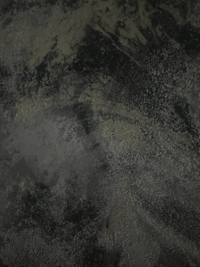 The image is a close-up, textured shot of a dark, patterned fabric. The texture appears to be a mix of raised dots and a subtle weave. The colors are predominantly dark grays and blacks, with some lighter, almost silver, highlights suggesting a slight sheen or different material interlaced. The lighting creates a sense of depth and highlights the uneven surface of the fabric. There are no people, objects, or any discernible elements that would indicate a specific location within Rosenovo, Bulgaria.