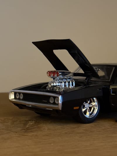 A black die-cast model of a Dodge Charger, likely a 1969 model, is positioned on a wooden surface. The hood is open, revealing a chrome engine with red intake runners. The front grille and headlights are visible, as is the driver's side front wheel with chrome rims. The background is a plain, neutral color. There is no indication of the city of Bruges, Belgium in the image.