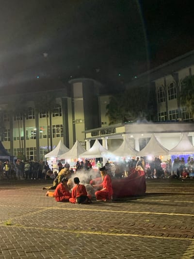The image was taken at night in front of a large building in the city of Kecamatan Batu, Indonesia. The building has several windows and a sign that reads "Graha Pancasila". Several white tents are set up in front of the building. There are many people in the area and some are standing around the tents. In the center of the image, four people are sitting on the ground in front of a large red blanket. There is smoke around them. One person is wearing a black cap and a red shirt, while the other three are all wearing red shirts. The people are sitting in a circle. The ground is paved with gray bricks. There is a large parking lot and many people in the distance. The image was taken on a clear night with no clouds in the sky. The area is lit by streetlights.