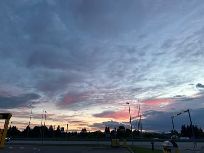 The image depicts a sunset over a semi-urban area in Laagri alevik, Estonia. The sky is filled with a dramatic display of clouds, ranging in color from deep gray and blue to vibrant pinks and oranges near the horizon, indicating the fading light of day. Several tall streetlights punctuate the scene, their dark silhouettes standing out against the colorful sky. A road runs horizontally across the middle of the frame, bordered by a row of trees that form a dark, dense line against the horizon. On the left side of the image, a yellow and black structure, possibly part of a bus stop or shelter, is visible. A car is partially visible on the road. Road signs and utility poles are also present in the background. The foreground consists of a paved surface, likely a parking area or sidewalk, with some low-lying green grass. The overall atmosphere is one of twilight tranquility, with the dramatic sky dominating the landscape.