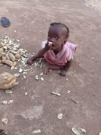 A young child is seated directly on a reddish-brown dirt surface in Unguwan Rimi, Nigeria. The child, with dark skin and dark hair styled close to the scalp, wears a pink sleeveless top with white ribbed trim. Its eyes are wide open, fixed on the viewer, and its mouth is open, containing a light-colored food item. The child's right hand holds this food to its mouth. To the left of the child, a cluster of unpeeled, light-brown potatoes and a pile of potato peels are present on the ground. Additional potato peels are scattered across the surrounding dirt. In the upper left background, a single dark-colored sandal rests on the ground.