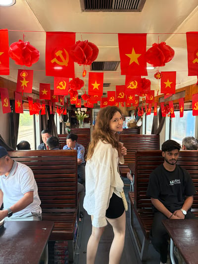 The photograph shows the interior of a train car in Huế, Vietnam. The car is adorned with strings of red Vietnamese flags and red paper lanterns. Several people are visible, seated in the car. A woman with long brown hair, wearing a white blouse, is smiling at the camera and holding a fan. A man wearing a black t-shirt sits across from her. Other people are seated further down the train car. The interior of the car is wood-paneled with wooden seating.