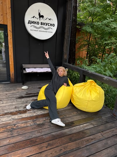 A Caucasian woman with blonde hair and fair skin is sitting on a large yellow beanbag chair outdoors. She is wearing a dark gray long-sleeved shirt and dark gray pants, with white sneakers. Her left arm is raised in the air, with her index and middle fingers forming a peace sign. She is looking to her left, with a neutral expression.

The beanbag chair is situated on a wooden deck. To the left of the woman, there is another yellow beanbag chair. Behind the beanbag chairs, there is a black wall with a round sign above it. The sign is white with black lettering and a silhouette of a deer and mountains. Text on the sign reads "ДИКО ВКУСНО" and "РЕСТОБАР ЛОКАЛЬНОЙ КУХНИ". Further to the left, a portion of a wooden structure and a glass door are visible. A wooden bench with a white cushion is positioned against the black wall. A single, light-colored stone is on the deck to the left of the woman.

To the right of the beanbag chairs, a dark wooden railing is visible, behind which lush green foliage and trees are present. The wooden deck appears to be slightly damp, with some areas showing reflections of water. The overall scene suggests an outdoor seating area of a restaurant or cafe. The location is identified as Nizhnekayancha, Russia.