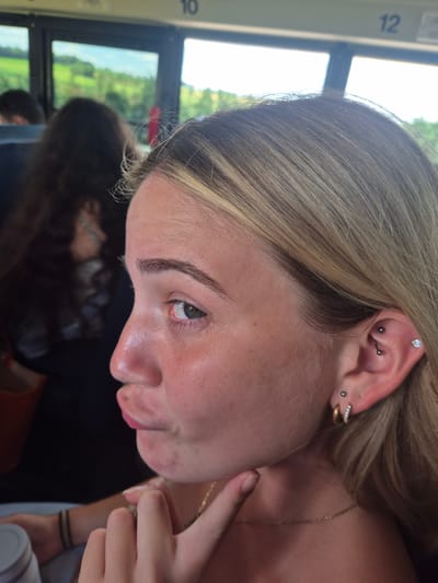 In a close-up, profile view, a young woman with blonde hair adorned with blonde highlights is captured. She is wearing multiple ear piercings: a small stud in her lower earlobe, a hoop earring with clear stones, and a curved barbell in her upper ear. Her skin is lightly freckled, and she has a light pink lipstick on her lips, which are puckered. Her left eye is visible, looking towards the left side of the frame. Her right hand is raised, with her index finger touching her chin. The background shows a bus interior with windows revealing a blurry green landscape under a bright sky. Two other individuals are visible in the background, also blurred. Numbers "10" and "12" are visible above the windows. The image is taken outdoors during the daytime.