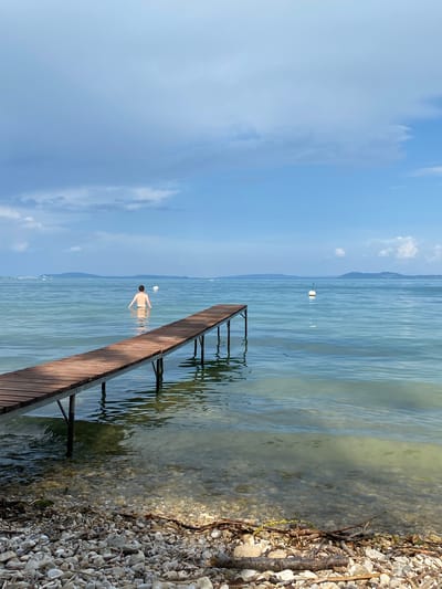 An unseen person stands in the clear, shallow water of a lake near the shore, facing away from the camera. The person is partially submerged, with the water reaching approximately to their waist. In the foreground, a wooden pier extends from the rocky shoreline into the water, casting reflections on the surface. The pier is constructed of horizontal wooden planks supported by dark metal legs. The lake water is calm, with visible ripples and a sandy or pebbly bottom apparent in the shallows. In the distance, across the expanse of water, a low, hazy mountain range forms the horizon under a partly cloudy sky. A few white buoys are visible floating on the water's surface. The environment is natural and serene, suggesting a lakeside setting.