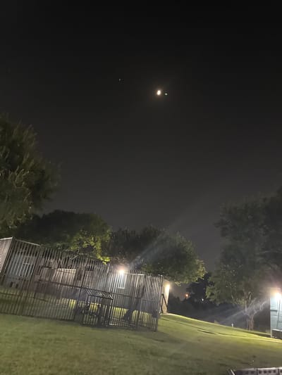 In the city of Dallas, United States of America, the image depicts a night scene. The sky is dark with some visible celestial bodies. In the upper portion of the image, two bright objects are visible, with one appearing larger and more luminous than the other, which is accompanied by a smaller, fainter point of light. Below the sky, trees with dense foliage are present, some illuminated by artificial light, casting shadows on the surrounding grass. In the foreground, a metal fence runs horizontally across the image, separating a grassy lawn from what appears to be a structure behind it, possibly a building with windows. The fence is made of vertical bars and has a gate-like structure within it. The ground is covered in grass, with subtle undulations. Artificial lights are visible, illuminating parts of the fence, the ground, and trees.