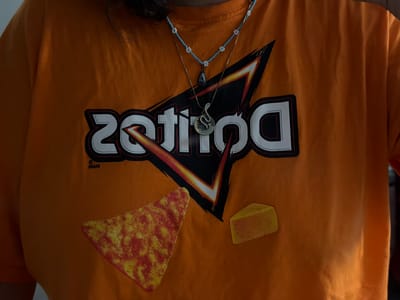 This is a close-up, head-on, medium shot of a person wearing an orange t-shirt with a Doritos logo and graphics of a Dorito chip and a slice of cheese. The t-shirt is the primary subject. The Doritos logo is in the center, with the brand name appearing to be backward, possibly as a stylistic choice. Above the logo, two necklaces are visible: one with blue beads and white flower charms, and another thinner chain with a pendant of a snake coiled around a moon. The graphic elements on the t-shirt include a large, stylized Doritos logo in black, white, and orange, and to the left and right of the logo, images of a cheese-flavored Dorito chip and a block of cheese. The background is the solid orange of the t-shirt, showing some texture and folds. There are no discernible elements that would place this image in Piacenza, Italy, or any other specific geographical location.