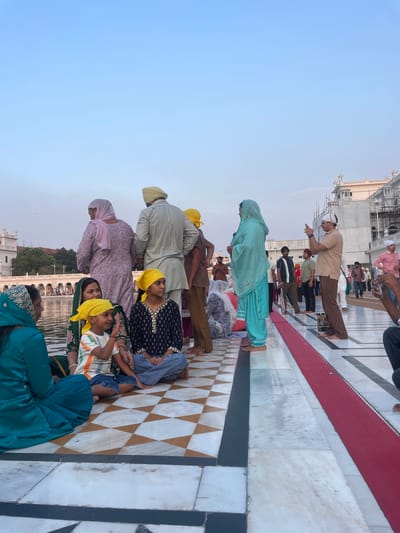 In Amritsar, India, at the Golden Temple complex, several people are gathered on a marble-tiled walkway next to a body of water. The individuals are dressed in traditional Sikh attire, with many wearing turbans and vibrant headscarves. A group of women and children are seated on the ground, some facing the water, while others engage in conversation. A man in the background is taking a photograph with his phone, and other visitors are walking along the pathway. The architecture of the Golden Temple, with its white marble and ornate details, is visible in the background, under a clear sky. The scene appears to be one of community and spiritual reflection.