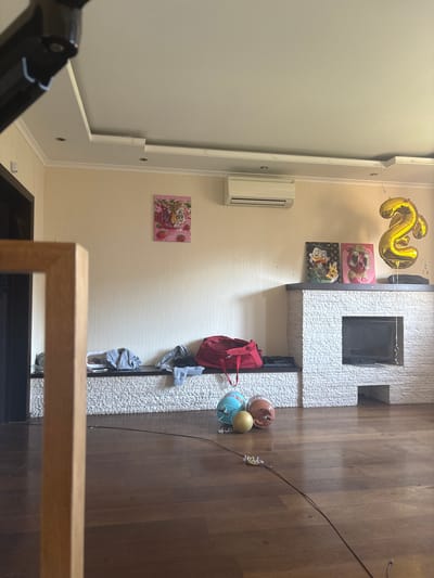 The image shows an indoor scene, likely a living room, in an apartment in Orekhovka, Russia. The room features a fireplace constructed with light-colored stone, a television set integrated into the fireplace, and two framed pictures above it. One picture depicts a cartoon character resembling Scrooge McDuck holding a dollar sign, and the other shows a cartoon bear wearing sunglasses and holding money. A large, gold-colored balloon shaped like the number "2" is tied to the fireplace mantel.
To the left of the fireplace, a long, bench-like shelf runs along the wall, decorated with a canvas painting of a tiger with strawberries. On this shelf, there are several pieces of folded clothing, including grey hoodies and dark-colored pants, as well as a red duffel bag.
In the foreground, on the wooden floor, are three spherical objects. Two appear to be decorative balls, one turquoise and the other peach-colored, and a smaller yellow sphere. A dark cable runs across the floor, connected to a small, tangled white object. A portion of a wooden picture frame is visible in the extreme left of the image, suggesting a photograph or artwork is being held or displayed nearby. The walls are painted a light, neutral color, and the ceiling has a modern design with recessed lighting. An air conditioning unit is mounted on the wall above the shelf. The overall impression is of a somewhat cluttered domestic space.