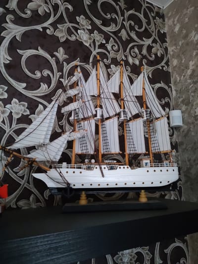 A close-up, eye-level, medium shot shows a detailed model ship on a black shelf. The ship is white with multiple sails, masts, and rigging. It is mounted on a black base with wooden stands. The background features dark brown wallpaper with a silver floral pattern and a textured grey wall on the right. A small white motion sensor is visible on the grey wall. The lighting is ambient, coming from the front and left of the scene. There are no people or animals present.