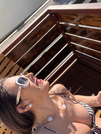 A woman is depicted relaxing outdoors on a bright, sunny day, captured from a high-angle perspective, head-on. She is lying on her back, looking upwards and slightly to the right, with her head tilted. She wears a black and white striped bikini top and a prominent gold-colored necklace featuring large, irregularly shaped white pearls. Matching pearl stud earrings adorn her ears. White-framed sunglasses, with the letters "DG" visible on the side arm, cover her eyes, reflecting the sunlight. Her lips are painted with red lipstick and are slightly parted in a relaxed expression. Her dark brown hair is visible around her head.
She appears to be resting on wooden furniture, likely a sun lounger, suggested by the wooden slats directly beneath her. Above her, a structure composed of dark wooden planks and lighter metal beams casts strong, striped shadows across the scene, indicating intense sunlight. A light-colored, pebbled or textured ground surface is visible in the lower left, consistent with an outdoor patio or poolside setting. The overall impression is one of leisure and enjoyment of warm weather. While no specific visual cues link the scene directly to Konz, Germany, the provided context identifies this as the location.