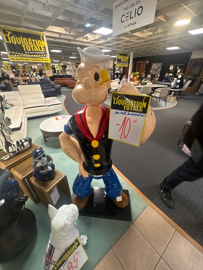 A large, colorful statue of Popeye the Sailor, featuring a light skin tone, white sailor hat with a yellow accent, black short-sleeved shirt with a red v-neck collar and yellow buttons, blue pants, and brown shoes, stands prominently in a furniture showroom in Schwindratzheim, France. The statue, depicted with a muscular right arm displaying an anchor tattoo, holds a yellow and white sign bearing the French text "LIQUIDATION TOTALE" (Total Liquidation) at the top, followed by handwritten "sur toute la durée" (for the entire duration) and a red "-10%". In front of Popeye, a white ceramic bulldog statue with blue and red stripes on its back holds a smaller sign displaying "LIQUIDATION" and "85". To the left, on a wooden display unit, a small blue and white skull-themed statue wearing a helmet is visible.
The spacious environment is brightly lit by overhead square fluorescent lights set into a grid ceiling. Multiple yellow banners advertising "LIQUIDATION TOTALE" and "Tout doit disparaître !" (Everything must go!) are suspended from the ceiling throughout the store. A large sign in the midground indicates "ESPACE CHAMBRE MEUBLES CELIO A L'ÉTAGE" (Bedroom Space CELIO Furniture Upstairs). The showroom floor transitions between light green, dark gray speckled carpet, and light brown tiles. Various furniture arrangements are displayed, including white and dark blue sectional sofas, brown leather sofas, and dining sets with tables and chairs. Several individuals, including adults and children, are present in the background, examining furniture. The lower legs and feet of a person wearing dark pants and black shoes are visible in motion on the right side of the foreground.