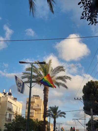 The image shows a view of the sky and urban landscape of Givatayim, Israel. Two flags are visible: one is the flag of Israel, and the other is a rainbow flag, commonly associated with LGBTQ+ pride. These flags are seen near palm trees and modern buildings, suggesting a public space. Power lines and streetlights are also present, indicating an urban environment. The sky is blue with scattered clouds, suggesting daytime. There are no people or specific actions depicted in the scene. The overall impression is of a city scene with symbolic flags displayed.
