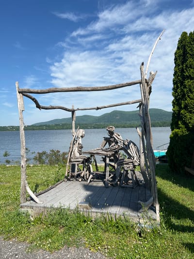 The image shows a rustic driftwood sculpture of two figures on a wooden deck overlooking a body of water, possibly the Hudson River. The deck is surrounded by grass and wildflowers. In the background, there are rolling hills and a blue sky with wispy clouds. The sculpture appears to be a stylized representation of two people seated or engaged in conversation. The location is likely near the waterfront in the Town of Fishkill, United States of America.