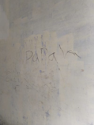 The image presents a detailed close-up of a textured wall surface in Keude Cunda, Indonesia. The wall exhibits a base color of light, desaturated beige, overlaid with irregular patches and vertical streaks of faded purplish-blue coloration. In the upper-central portion of the wall, the word "PaPaH" is handwritten in dark, likely black, script. Below and slightly to the left of this primary inscription, fainter and thinner dark scribbles are visible, including partially discernible text that appears to read "Panah Cunda." The wall surface displays signs of wear and a granular texture. No individuals, additional objects, or specific interactions are depicted within the frame.