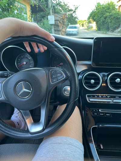 An inside view of a Mercedes-Benz C-Class W205, captured from the driver's perspective, shows a hand resting on the steering wheel. The car is parked on a dirt road in General Todorov, Bulgaria, with a silver Volkswagen Golf visible in the distance. The interior features a black dashboard with a central infotainment screen, dual air vents, and various control buttons. A phone is charging in a designated slot below the climate controls. The exterior view, seen through the windshield, reveals a rural setting with houses and trees. The image is taken in daylight.