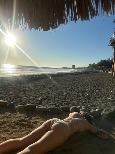 A woman in a light-colored bikini is lying on a dark sandy beach. She rests near a row of rocks or boulders that separate the sand from the beach's edge. In the background, a body of water meets the horizon under a clear blue sky. A bright sun is visible, and the light creates a glare on the water. Part of a thatched roof is visible in the corner.