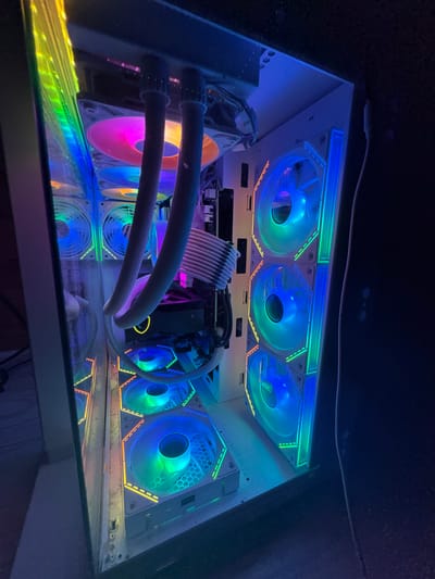 In a dimly lit room, a custom-built computer tower case is illuminated by an array of RGB lights. The case is white and has a transparent side panel, revealing the internal components. Several large, circular fans with RGB lighting are visible, arranged vertically along the front and horizontally at the top and bottom of the case. These fans display a gradient of colors, shifting from pink and orange at the top to blue and green towards the bottom. Two thick, gray hoses, likely part of a liquid cooling system, extend from the top of the case. A graphics card is partially visible, also adorned with RGB lighting. The overall impression is one of a high-performance gaming or workstation computer with a visually striking aesthetic. The image was taken in Busmantsi, Bulgaria.