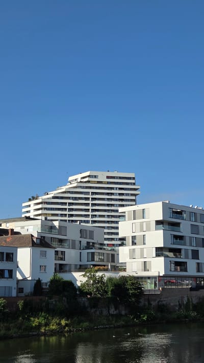 The image depicts a modern architectural complex situated along a river in Ulm, Germany. The composition features a cluster of white buildings with sharp geometric lines, characteristic of contemporary design. A prominent, multi-story building with a stepped, tiered facade dominates the background, its numerous balconies and windows reflecting the clear blue sky. In the foreground, a lower, also white, building with balconies and glass railings stands. The river in the foreground is dark and reflects the buildings and the lush greenery along its banks. There are no people visible in the image, but two ducks are present on the water. The overall scene is one of urban development integrated with a natural setting.