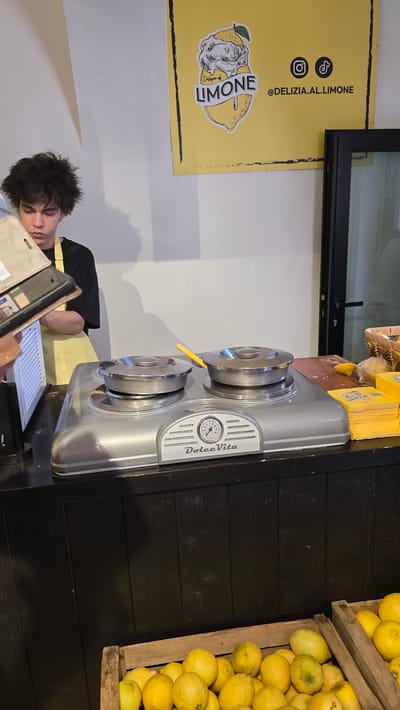 This is a medium shot, taken outdoors in the daytime. The scene depicts a street vendor in Limone sul Garda, Italy. A young person with dark, messy hair is slumped over a counter, seemingly asleep or exhausted. They are wearing a black t-shirt and a yellow apron. In front of them is a tablet or e-reader.

To the left of the person is a bright yellow sign with black text and graphics. The sign features a logo with a drawing of a lemon and a classical bust, and text that reads "LIMONE" and "@DELIZIA.AL.LIMONE." There are also social media icons for Instagram and TikTok.

To the right of the person is a shiny, silver appliance with two circular stovetop-like elements and a gauge that reads "Dolce Vita." A bright yellow utensil is resting on one of the elements. Next to the appliance is a wooden crate overflowing with fresh, yellow lemons. The background consists of a dark wooden wall and a white wall. The overall impression is a casual, perhaps late-night or early-morning, scene at a lemon-themed vendor stall.