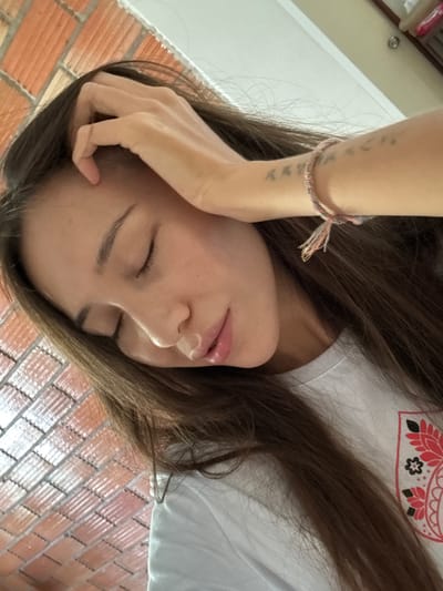 In a close-up, overhead shot, a young woman with long, dark brown hair is seen with her eyes closed and her head tilted to the side, resting on her hand. Her hair falls across her face and shoulders. She is wearing a white t-shirt with a circular, red and black patterned emblem on the left side of her chest. A thin, woven bracelet with multicolored threads is visible on her left wrist, and a faint tattoo is present on her forearm. The background consists of a textured, brick-like pattern in shades of orange and white. The image appears to be taken indoors, possibly in the city of Baan Nok Sai, Thailand, given the lighting and the architectural hint of the background.