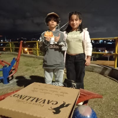 Two children stand outdoors near playground equipment at night. A girl, taller, wears a light jacket and dark pants. A boy, holding a slice of pizza, gives a thumbs-up. A pizza box with "Cavallino" branding sits nearby. City lights twinkle in the background beneath a dark sky. The scene appears relaxed and casual.