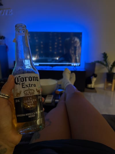A person is holding a bottle of Corona Extra beer, which is partially filled. The background shows a television screen displaying a bright blue light and some abstract shapes. To the right of the TV, a person's legs are visible, wearing white socks. The setting appears to be indoors, possibly a living room, with soft lighting. The overall mood is relaxed and casual. The visible text on the bottle reads "Corona Extra," "LA CERVEZA MAS FINA," "CERVECERIA MODELO, S. DE R.L. DE C.V.," "MEXICO," and "330 ml." The time of day cannot be determined from the image.